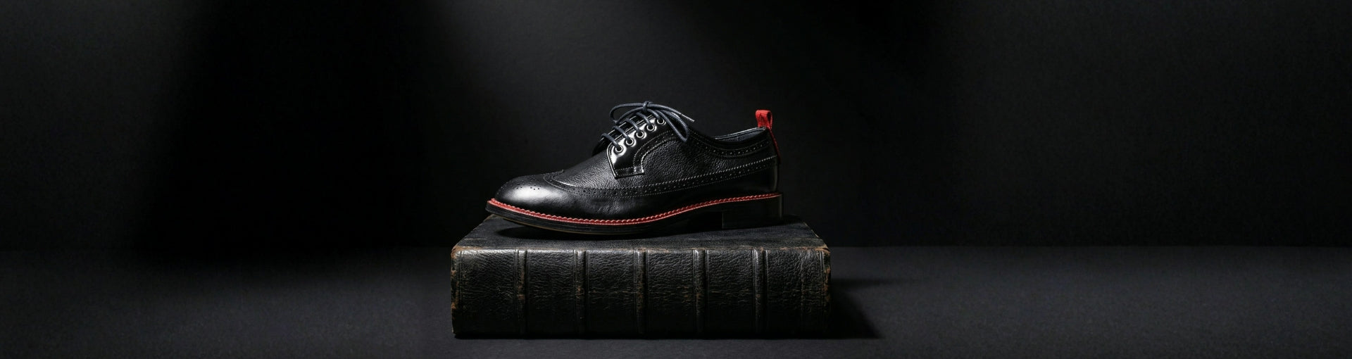Handmade black leather oxford shoe with signature red welt placed on a vintage book, representing traditional European shoemaking craftsmanship.